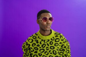 A studio portrait shot of a young man wearing a leopard print jumper and sunglasses, standing in front of a purple backdrop and looking off to the side with a contented look on his face.