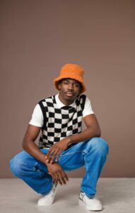 Portrait of fashionable young man wearing checker chess sweater vest blue jacket, jeans pants and orange bucket hat looking at camera. Studio shot on brown background.