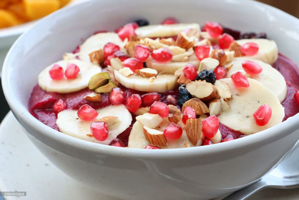 Stock photo showing a healthy eating breakfast fruit bowl of beetroot and strawberry smoothie puree, sliced banana, pomegranate seeds, blueberries and chopped almonds on al fresco beach restaurant table.